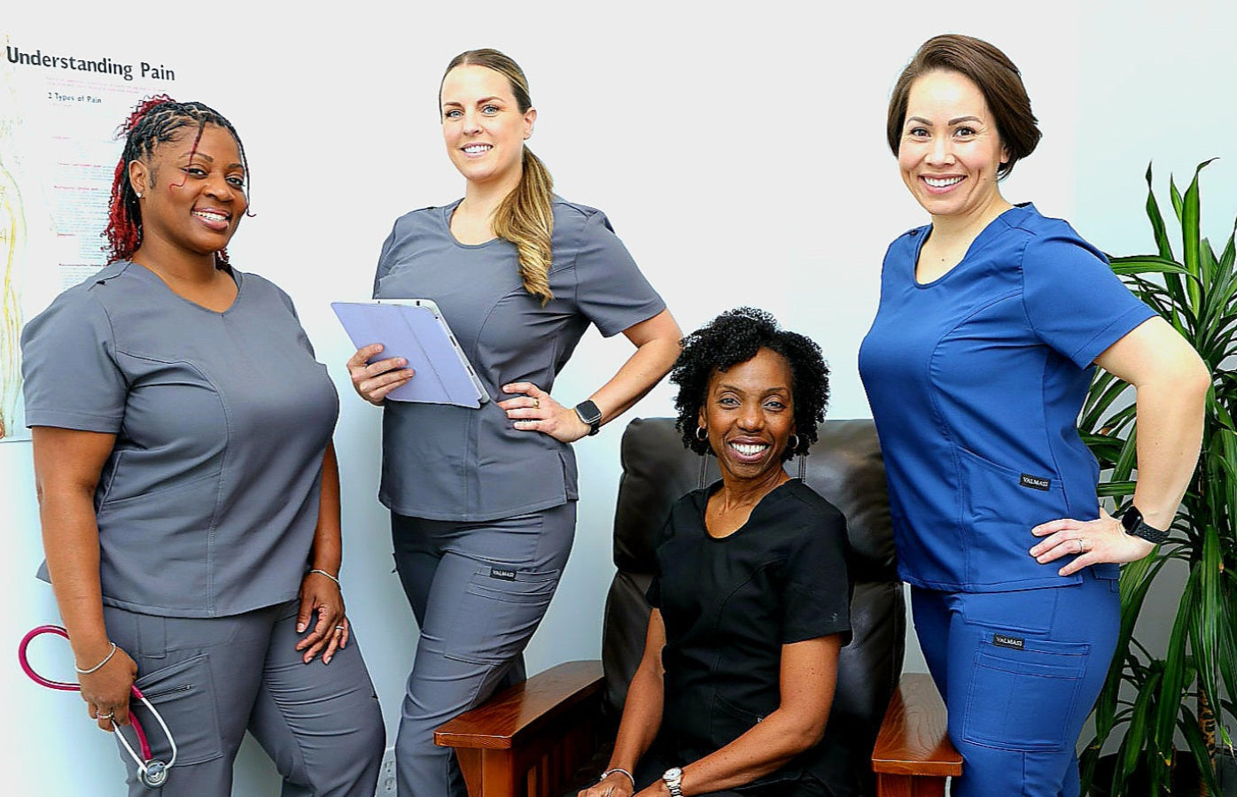 Four healthcare professionals in various scrubs posing together in a room with a plant.